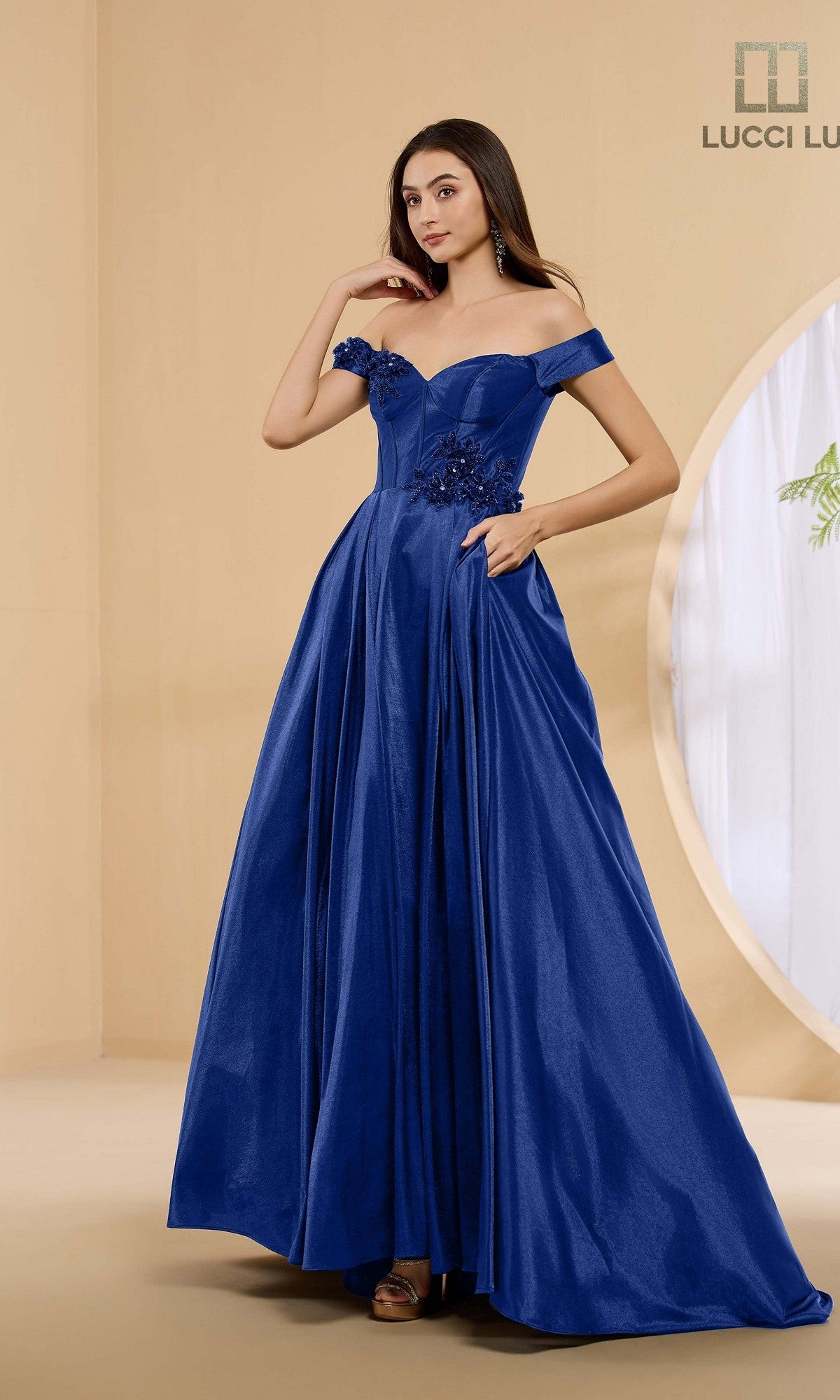A woman poses in a Lucci Lu 1472 long prom dress in midnight blue, featuring an off-the-shoulder a-line silhouette, floral appliques, and pockets, standing in a minimalist room with beige walls.